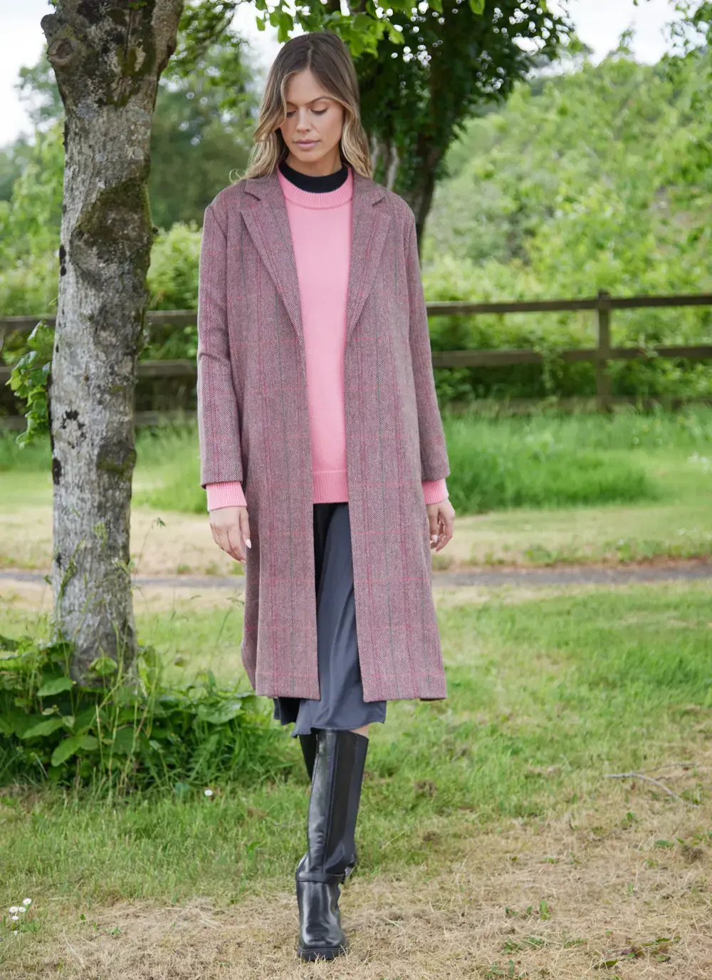 Woman wearing a brown and pink herringbone Irish tweed duster coat with lapel collar, styled over a pink sweater and black boots.
